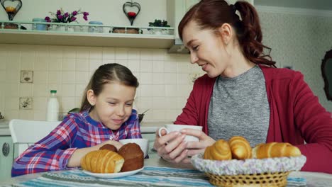 Cheerful-mother-and-cute-daughter-having-breakfast-eating-muffins-and-croissants-talking-at-home-in-modern-kitchen.-Family,-food,-home-and-people-concept