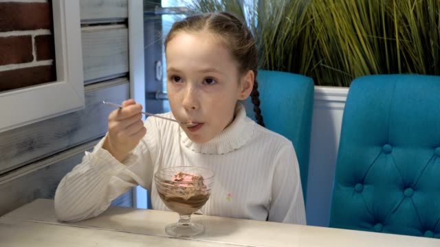 A-cute-little-girl-with-freckles-on-her-face-in-a-white-dress-is-sitting-in-a-cafe-and-eating-strawberry-ice-cream-with-chocolate.-Portrait.-High-detail.-4K.-25-fps.