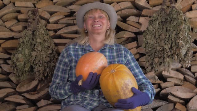 Happy-Woman-On-A-Background-Of-Stacked-Firewood-Holding-In-Her-Hand-Ripe-Pumpkin