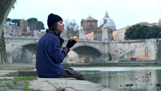 young-homeless-lonely-woman-sitting-at-the-river-eats-an-apple