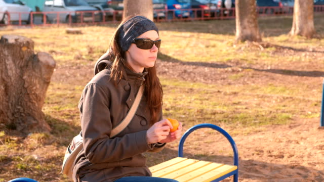 Young-brunette-woman-in-sunglasses-peels-and-eats-tangerine-sitting-on-the-bench-in-park.