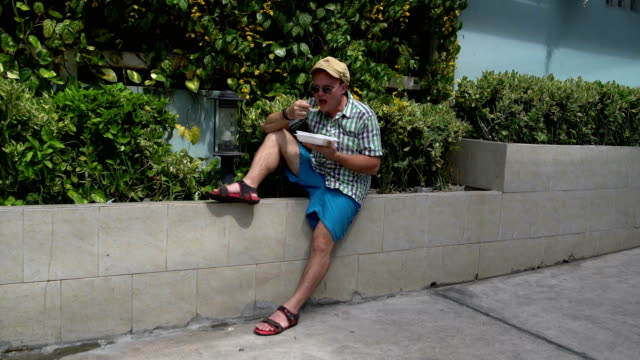 Street-food.-A-man-sitting-on-the-street-on-a-parapet-eating-stewed-vegetables-from-a-container