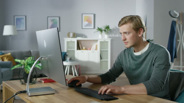 Portrait-of-the-Focused-Young-Man-Working-on-a-Personal-Computer-while-Sitting-at-His-Desk.-In-the-Background-Cozy-Living-Room.