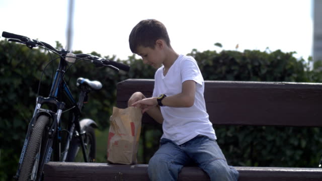 Junge-sitzt-auf-einer-Bank-in-einem-Park-öffnet-ein-Paket-und-nimmt-heraus-essen,-ein-Picknick