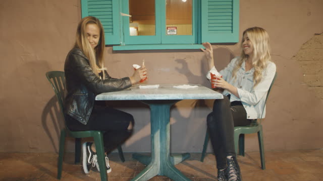 Two-girls-sitting-at-table-outside-playing-with-food-throwing-it-at-each-other-laughing