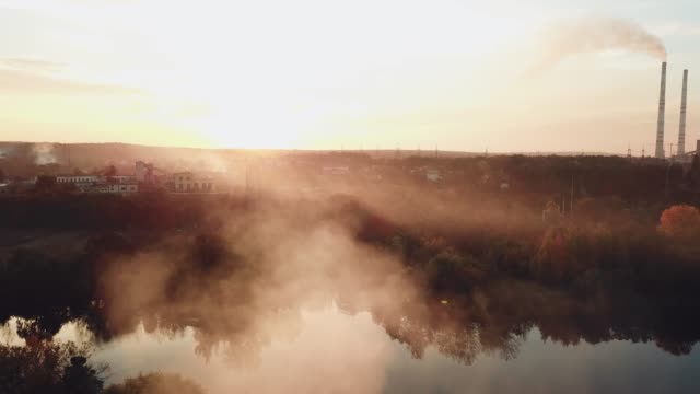 Rauch-vom-Feuer-breiten-sich-über-den-Fluss-bei-Sonnenuntergang-auf-dem-Hintergrund-eines-Kraftwerks-mit-zwei-Rohren.