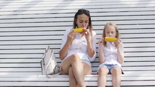 Young-mother-with-her-daughter-eating-boiled-corn-in-the-park-in-summer.