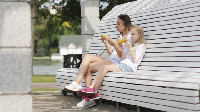 Una-mujer-joven-con-una-chica-rubia-y-descansar-en-el-parque-y-comer-maíz-caliente.