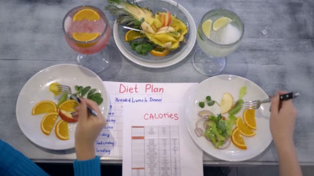 comida-vegana-saludable,-chicas-comen-fruta-de-placas-sentado-en-la-mesa-con-plan-de-dieta-y-tabla-de-calorías