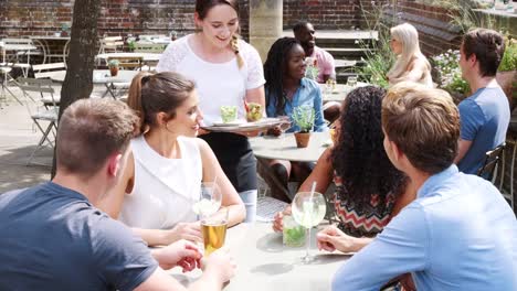Grupo-de-reunión-de-amigos-para-las-bebidas-en-las-mesas-al-aire-libre-en-restaurante-ser-sirve-bocados-por-camarera