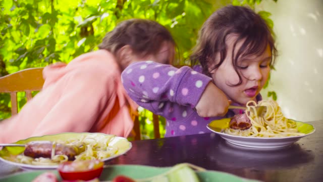 Zwei-freche-Mädchen-am-Tisch-sitzen-und-Essen-Pasta-und-Bratwurst