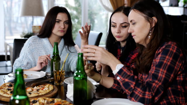 Alegre-compañía-de-las-niñas-en-el-restaurante-viendo-fotos-en-el-smartphone.-Chicas-comer-pizza,-beber-cerveza,-charlar-y-divertirse.