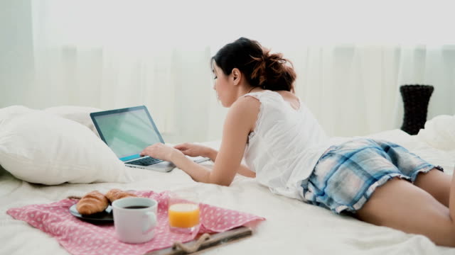 Young-woman-using-laptop-during-breakfast-lying-on-white-bed-at-home.-Brunette-girl-typing-on-pc-computer