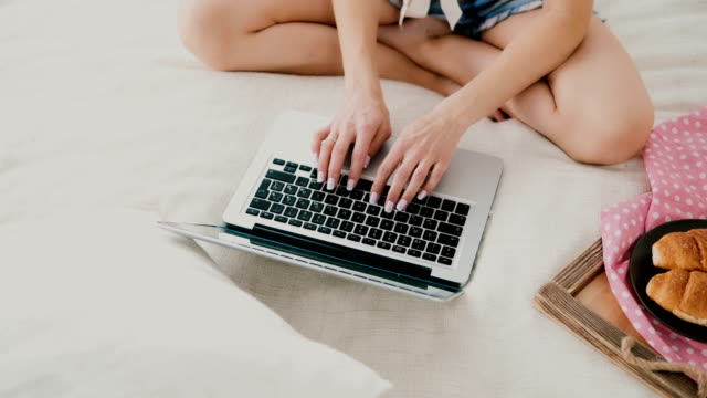 Young-woman-using-laptop-during-breakfast-sitting-on-white-bed-at-home.-Close-up-view-of-girl-typing-on-pc-computer