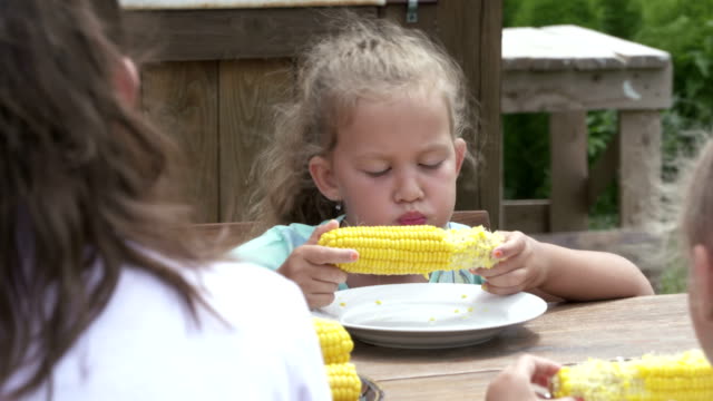 Kleines-Mädchen-isst-Mais-Maiskolben-auf-dem-Esstisch-im-freien