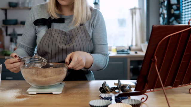 Young-beautiful-woman-mixing-ingredients-in-bowl,-using-whisk.-Attractive-female-looking-at-recipe-from-cooking-book