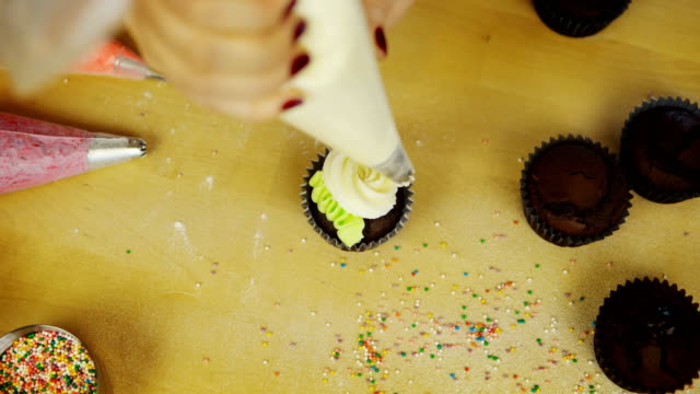 Vista-superior-de-las-manos-del-joven-decorando-los-cupcakes-de-chocolate-con-crema-de-colores.-Mujer-usando-la-Manga-pastelera