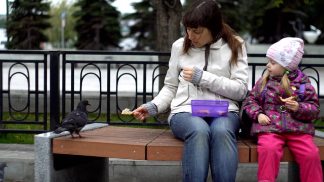 Mother-and-daughter-eat-sandwiches-and-feed-pigeons-on-a-cold,-cloudy-day.