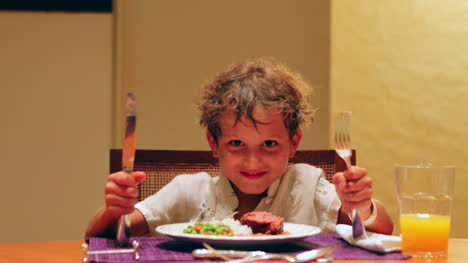 Niño-esperando-comida-llegar-a-la-mesa-en-4K-niño-joven-con-tenedor-y-cuchillo-en-la-mesa-pidiendo-comida-riendo-y-sonriendo-a-la-cámara-en-4K