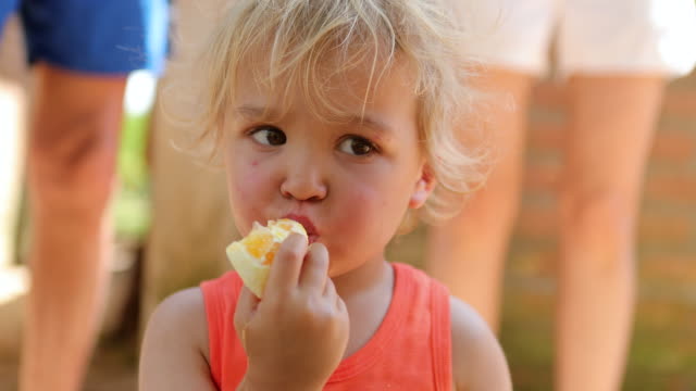 Porträt-von-cute-blonde-Kleinkind-Jungen-essen-eine-Orange-in-4K.-Reportageaufnahme-des-jungen-Säuglings-beiläufig-essen-gesundes-Obst-draußen-im-Sonnenlicht-in-4K-60fps