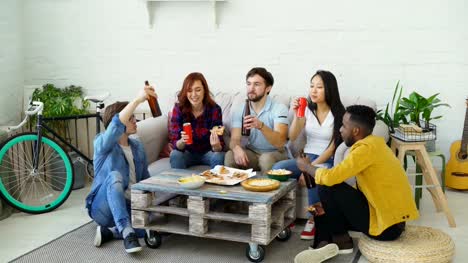 Glückliche-Freunde-Pizza-essen-und-feiern-party-klirrende-Flaschen-mit-Bier-und-Limonade-sitzen-zu-Hause-auf-sofa