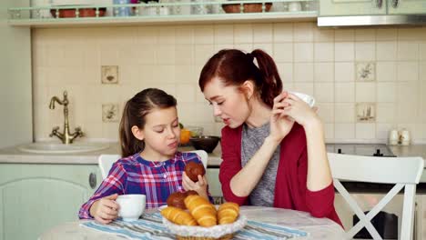 Happy-mother-and-cute-daughter-having-breakfast-eating-muffins-and-talking-at-home-in-modern-kitchen.-Family,-food,-home-and-people-concept