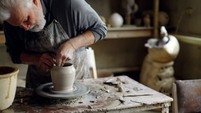 Experienced-ceramist-grey-haired-bearded-man-is-smoothing-molded-ceramic-pot-with-wet-sponge.-Spinning-throwing-wheel,-muddy-work-table-and-clayware-are-visible.