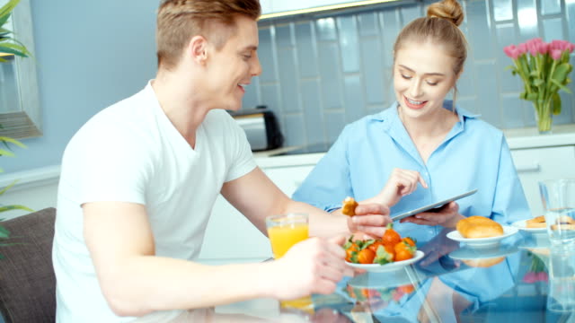 Young-couple-using-digital-tablet-while-having-breakfast-at-kitchen-table.