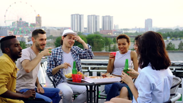 Multiracial-group-of-friends-is-clinking-glasses,-drinking-soft-drinks-and-eating-pizza-sitting-at-table-on-rooftop-celebrating-friends-reunion.-Food-and-party-concept.