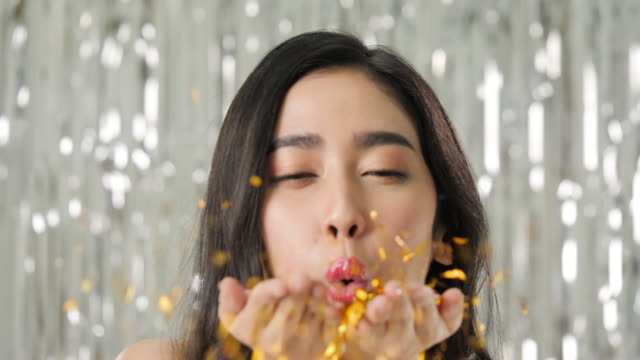 Schöne-asiatische-Frau-bläst-gold-Glitter-Konfetti-oder-Stücke-von-Goldfolie-vor-glänzenden-Schimmer-Vorhang-Hintergrund,-Slow-motion