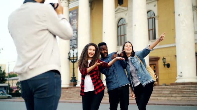 Professional-photographer-is-shooting-group-of-travelers-happy-young-people-posing-for-camera-in-front-of-beautiful-theater-with-backpacks-and-laughing.