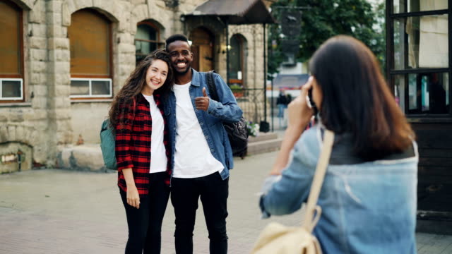 Young-woman-is-taking-pictures-of-beautiful-couple-African-American-guy-and-Caucasian-girl-standing-in-the-street-and-posing-for-camera.-Tourism,-technology-and-friends-concept.