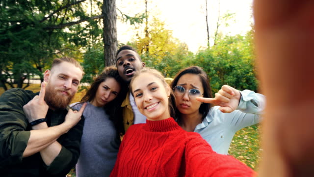 Sicht-der-jungen-Frau-Haltevorrichtung-mit-Kamera-und-Selfie-mit-multi-ethnischen-Freundesgruppe-im-Park-im-Herbst-gedreht.-Konzept,-Fotografie-und-Menschen.