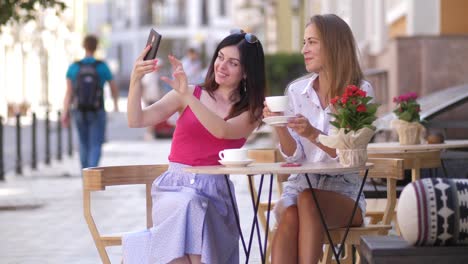 Two-beautiful-young-women-sit-at-a-table-in-a-cafe-and-take-a-selfie-on-the-phone-4k-slow-motion