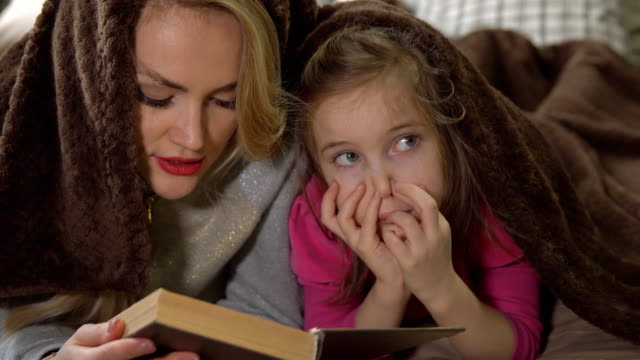 Mother-and-daughter-lie-on-a-bed-under-a-blanket-and-read-a-book.