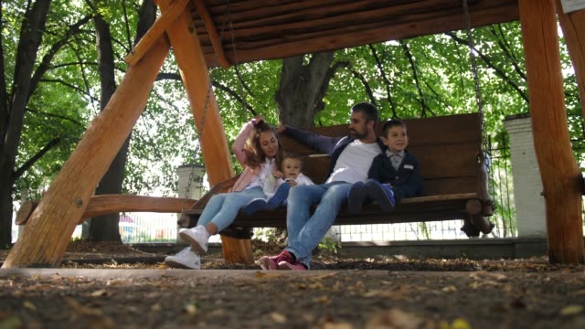Glückliche-junge-Familie-Vater,-Mutter,-Sohn-und-Töchterchen-reitet-auf-einer-Schaukel-im-Park.-Lachen-und-Lächeln.-4-k.-Slow-Motion