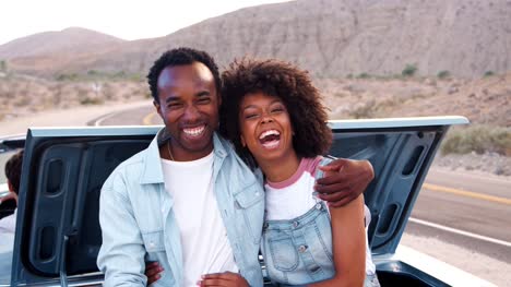 Laughing-couple-sitting-on-car-at-roadside-stop-on-road-trip