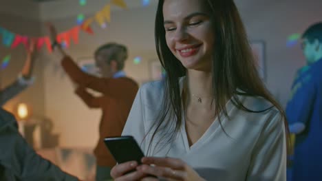 At-the-Wild-House-Party:-Beautiful-Young-Girl-Uses-Smartphone,-Browses-Through-Internet-and-Social-Network.-In-the-Background-Crowd-of-Young-People-Dancing-Off-and-Having-Fun.