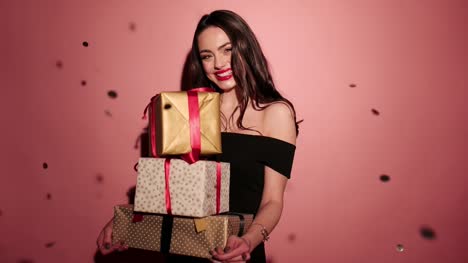 Brünette-Frau-glücklich-halten-Sie-ihr-Geschenke-mit-Konfetti-in-rosa-Hintergrund-schwarz-Kleid-tragen