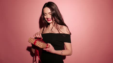 Brünette-glückliche-Frau-öffnen-Sie-ihr-Geschenk-mit-Konfetti-in-rosa-Hintergrund-tragen-schwarze-Kleid