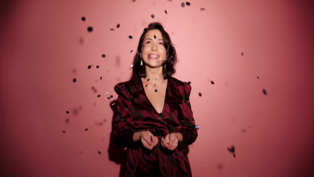 Brunette-happy-woman-throw-up-and-laugh-to-camera-with-confetti-in-pink-background-wear-red-dress