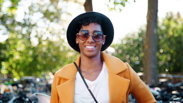 Fashionable-young-black-woman-standing-outdoors-wearing-sunglasses,-a-yellow-coat-and-a-black-hat-looking-to-camera-and-laughing,-close-up,-focus-on-foreground
