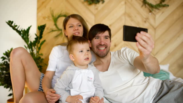 Lächelnde-Eltern-mit-Baby-nehmen-Selfie-Familienfoto-auf-Bett-zu-Hause
