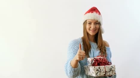 Glückliches-Mädchen-in-die-Nikolausmütze-und-mit-Geschenken-einen-Daumen-auftauchen-auf-weißem-Hintergrund