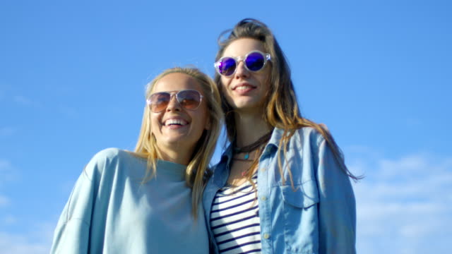 Dos-hermosas-chicas-felizes-llevando-gafas-de-sol-abrazan,-ríen-y-tienen-un-montón-de-diversión.-Cielo-azul-de-fondo.