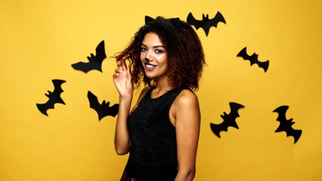 Ziemlich-Afro-Amerikanerin-mit-Katze-Make-up-und-Ohren-Lachen-und-flirten,-Kamera-über-gelbe-Wand-mit-Fledermäusen-isoliert