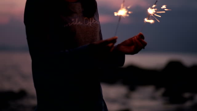 Young-asian-woman-stands-on-beach-with-sparkler-at-sunset-in-slow-motion.