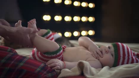 Cute-baby-boy-dressed-in-striped-christmas-clothes-is-lying-on-beige-bed-and-smiling-while-mother-carefully-holding-his-feet