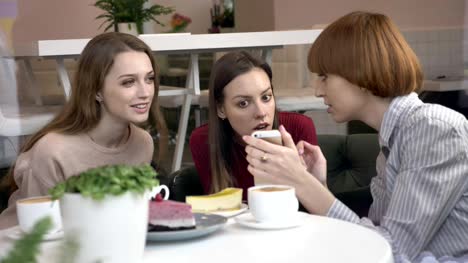Three-young-caucasian-girls-are-sitting-in-a-cafe,-laughing,-smiling,-friends,-company,-gossips,-dialogue,-discussion.-Girlfriends-in-the-cafe-concept.-Using-a-smartphone,-watch-a-funny-video,-a-photo,-shocked-60-fps