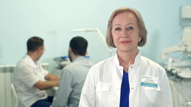 Senior-female-doctor-looking-at-camera-while-male-doctor-talking-to-patient-on-the-background